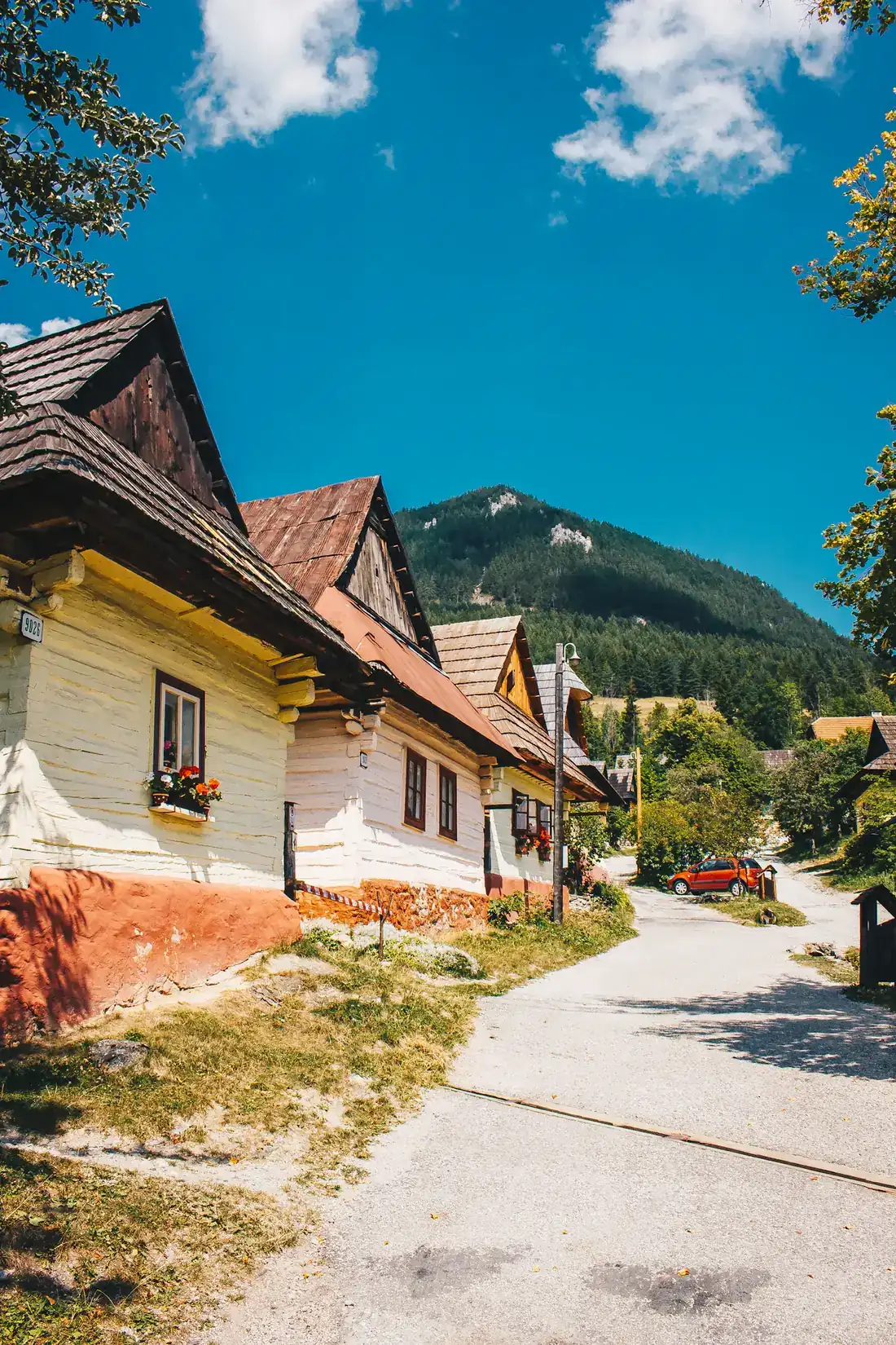 Ulica s tradičnými drevenými domami s bielymi stenami, v pozadí zalesnený vrch a modré nebo